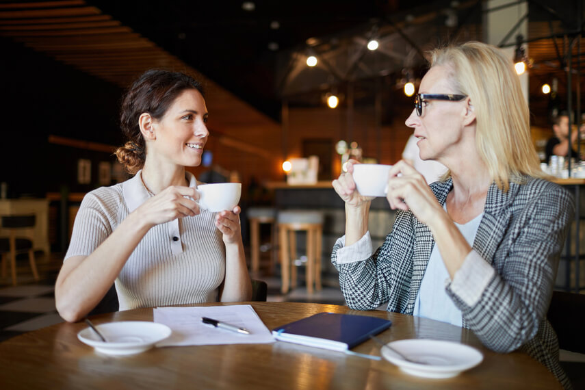 Two women drink coffee and talk at a coffee shop, with a pen and notebook on the table between them